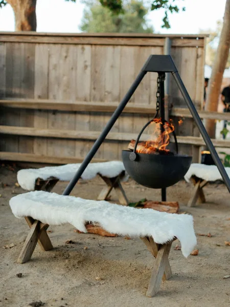 Fire pit lounge outdoor wedding lounge with a hanging bowl on tripod, firewood and benches topped with white faux fur throws by a wood fence