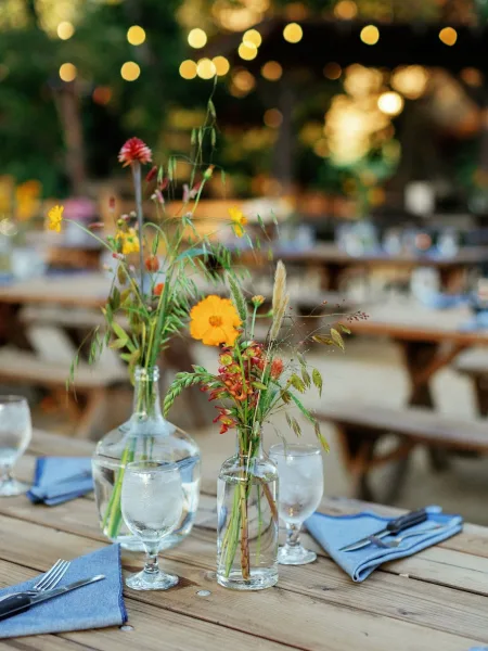 Reception tablescape with wildflower wedding centerpieces in glass vases, blue linen napkins and water goblets on long wooden tables under string lights outdoors