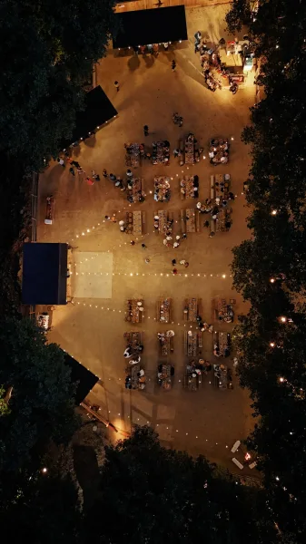 Outdoor reception layout with aerial wedding reception view of banquet tables under string lights, tent canopies and lounge seating in a tree-lined courtyard at night