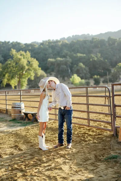Couple kiss portrait of a cowboy wedding couple holding hands by a metal gate fence, bride in short white dress and boots, both in cowboy hats