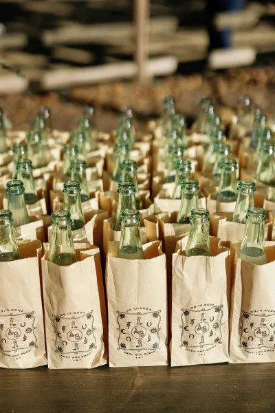 Wedding welcome bags with hotel welcome bags wedding labels, glass bottle favors, and monogram date tags stacked on a wood table indoors