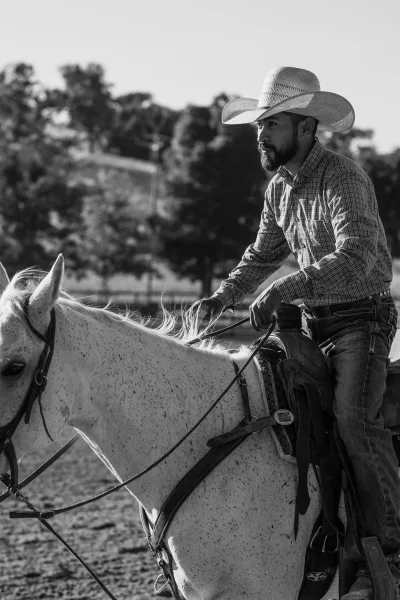 Cowboy on horseback in a western engagement photo, wearing a cowboy hat and plaid shirt, riding through a fenced pasture with trees and hillside