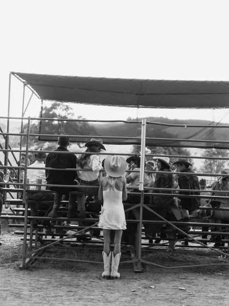 Wedding guest candid of western wedding guests in cowboy hats and boots on shaded bleachers, sipping wine beside bottles in a field with hills