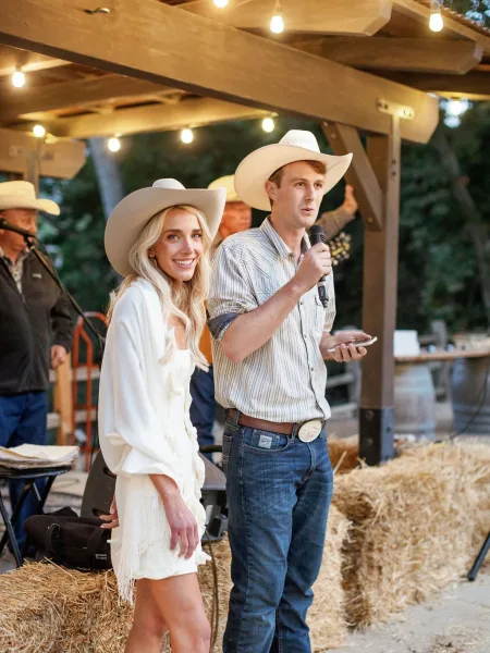 Wedding speech with cowboy hats as a guest speaks into a handheld microphone under string lights in a wood pavilion with trees and musicians