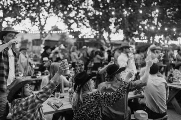 Wedding reception toast as guests raise glasses under string lights, with cowboy hats visible at wooden picnic tables among trees