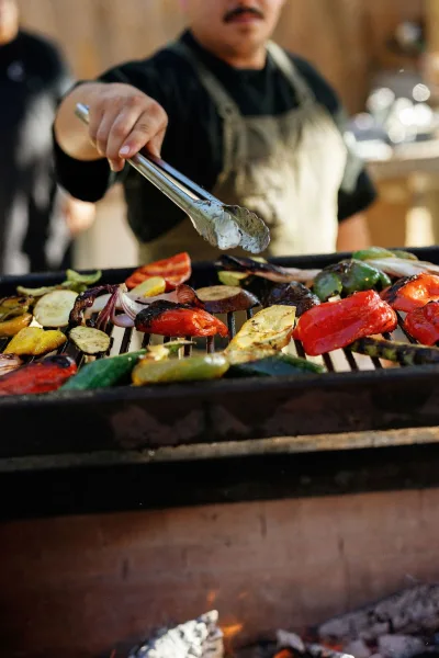 Wedding catering at a live cooking station as a chef uses metal tongs to grill vegetables at an outdoor setup with blurred guests and warm light