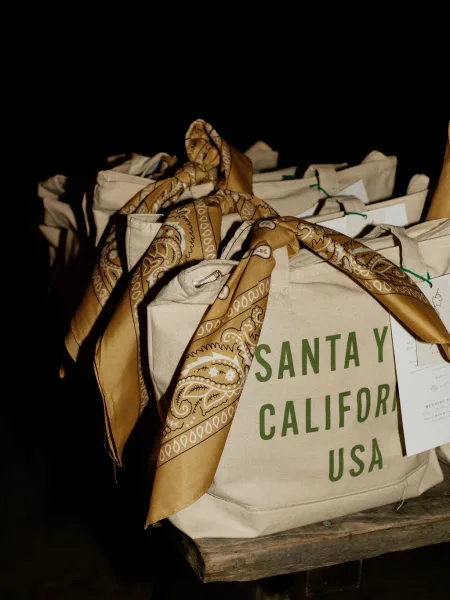 Wedding welcome bags displayed as canvas tote welcome bags with bandana scarves, luggage tags, and paper cards on a wood table against a dark backdrop