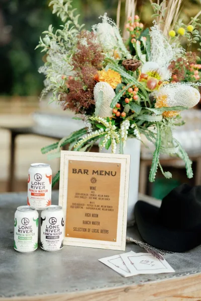 Wedding bar menu and signature drinks sign in a framed display on a bar counter, with a floral arrangement in a ceramic vase and canned drinks