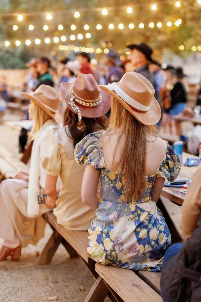 Wedding reception guests in cowboy hats mingle in floral dresses around wooden picnic tables under glowing string lights at dusk