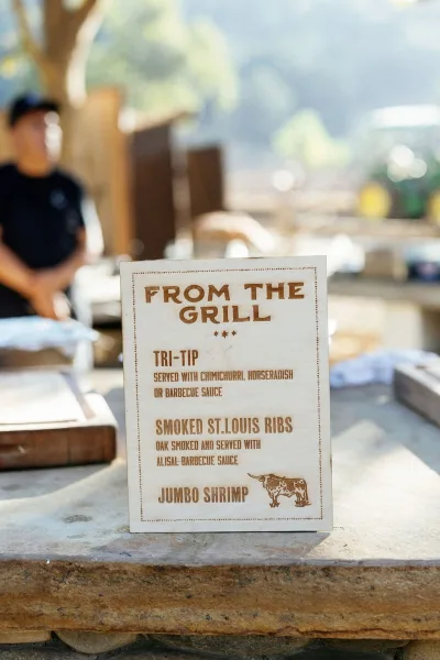Wedding food sign listing the menu on a bar counter, printed card at an outdoor catering station with hills and guests behind