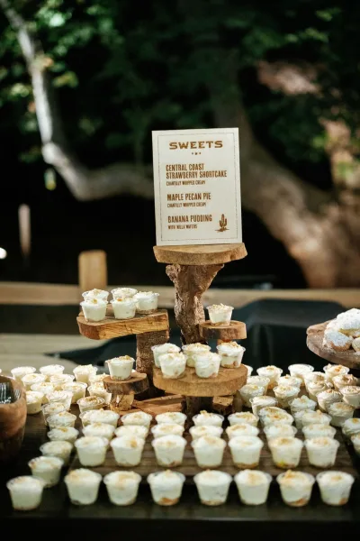 Wedding dessert table with rustic dessert display of mini dessert cups, wooden tiered stands and a dessert menu sign set by trees