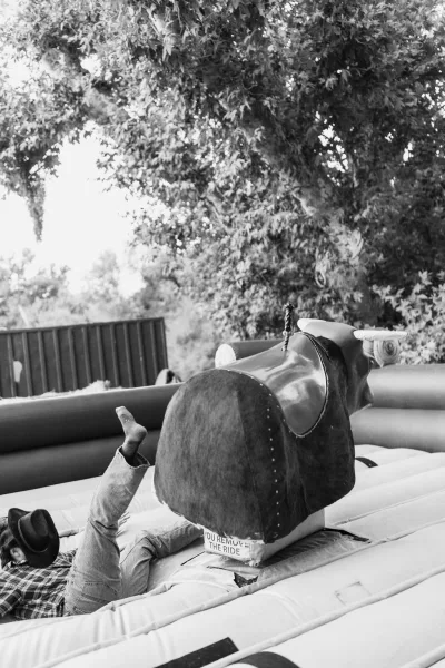 Mechanical bull ride at a wedding mechanical bull setup, guest in cowboy hat and plaid shirt on inflatable bull by trees and fence