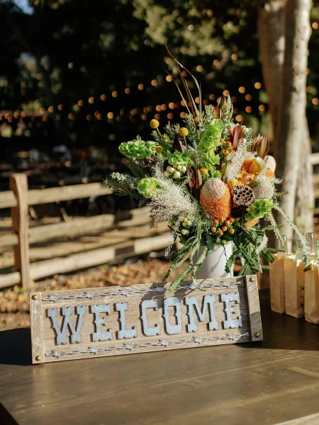 Wedding welcome sign on a wooden table with metal letters and a floral vase with greenery and berries beneath string lights in trees outdoors