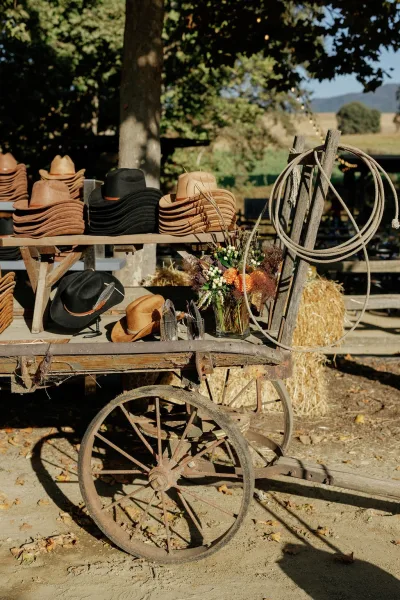 Wedding welcome table rustic wedding welcome display with cowboy hats on a wooden wagon, hay bales, flowers, and string lights in a field