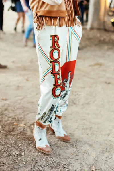 Wedding guest outfit with western wedding guest outfit vibes, pairing a fringe jacket, printed pants, and cowboy boots on dirt ground by a blurred crowd