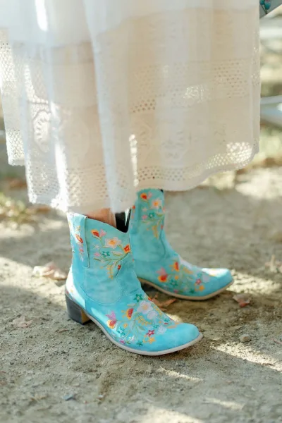Wedding boots in turquoise cowboy boots with floral embroidery peeking beneath a lace dress hem on dirt ground with scattered leaves