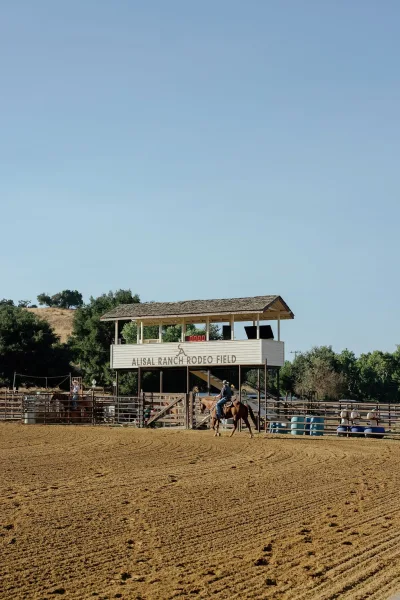 Horseback riding in a rodeo arena as a cowboy hat rider guides a horse past barrels and fencing under a blue sky with hills