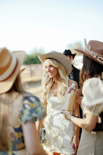 Bride portrait of a cowgirl bride in a short wedding dress with a cowboy hat, holding a clutch outdoors by a wooden fence and trees