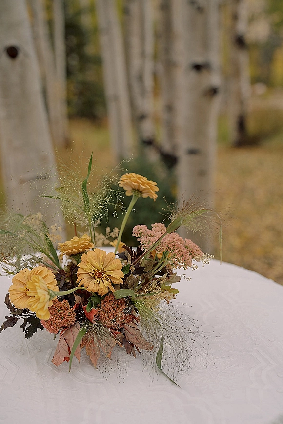 Bridal bouquet with yellow bridal bouquet blooms and greenery on a white lace tablecloth, set against birch trees and fall foliage