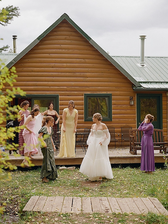 Bridal party getting ready as bride with bridesmaids in pastel dresses gather on a log cabin porch beside outdoor chairs under gray skies