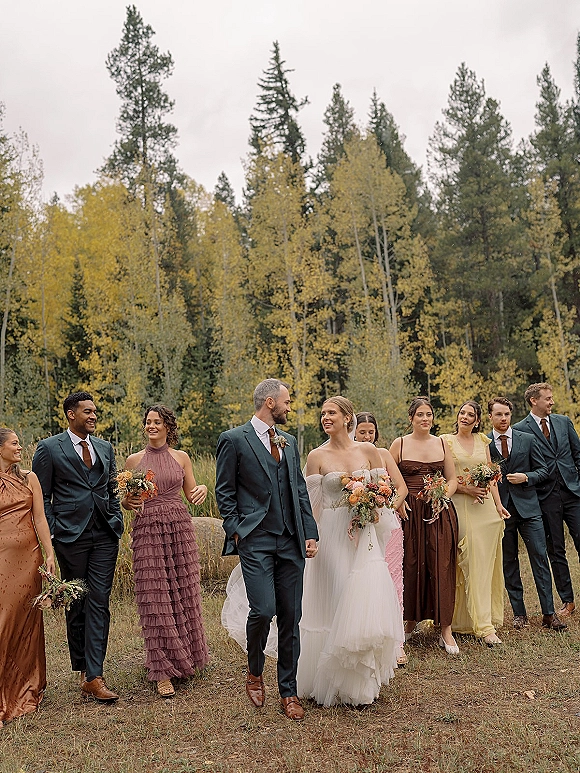 Wedding party portrait with bride and groom with wedding party walking across a grassy field, bouquet accents, autumn trees under overcast sky