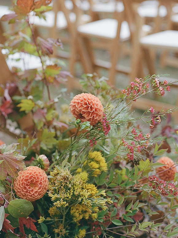 Wedding florals with autumn wedding flowers, dahlias and pink waxflower with greenery and autumn leaves beside wooden chairs indoors