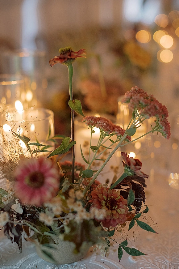 Wedding centerpiece with floral arrangement and candles in glass holders on a white tablecloth, with bokeh reception lights behind