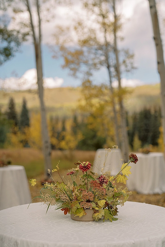 Wedding centerpiece with wildflower wedding centerpiece accents, greenery and autumn leaves in a low bowl on a round linen table in a mountain meadow reception