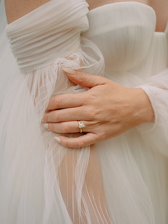 Engagement ring with an emerald cut diamond on a gold band, worn on a manicured hand beside a tulle bridal gown sleeve and ruched bodice