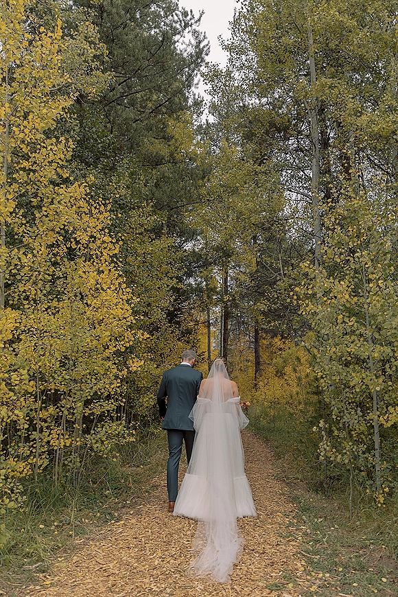Couple portrait of bride and groom walking away down a woodland path, her veil and dress train flowing beside autumn trees and leaves