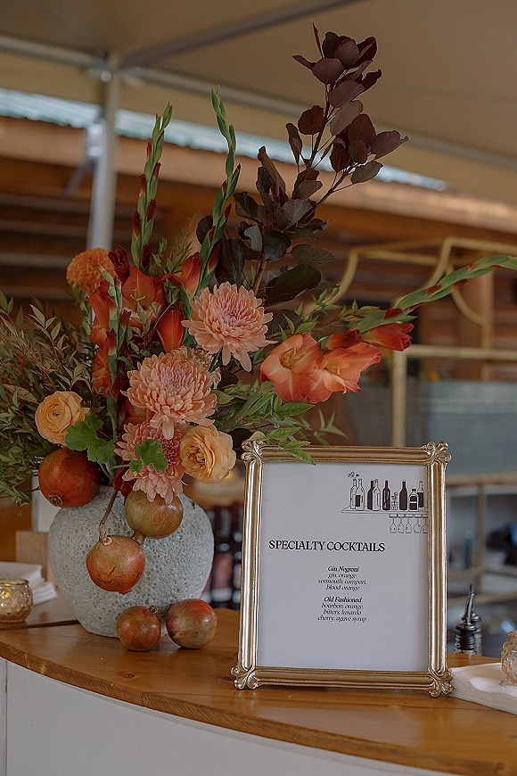 Cocktail bar signage in a gold frame on the wedding counter, paired with a floral arrangement in a ceramic vase and pomegranates under a tent canopy