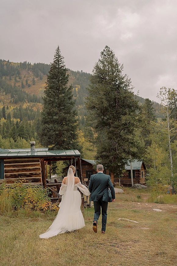 Couple walking away, bride and groom walking with long veil and off-the-shoulder gown on a grassy path by a log cabin and pines, mountains beyond