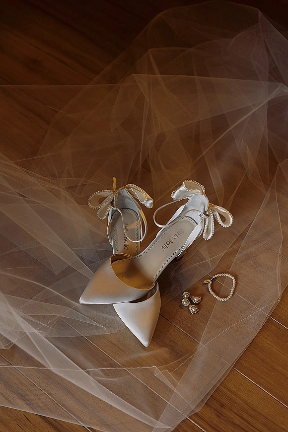 Bridal shoes in white wedding heels with ankle bows and pearl trim, styled on a tulle veil with pearl jewelry on a wood floor flatlay