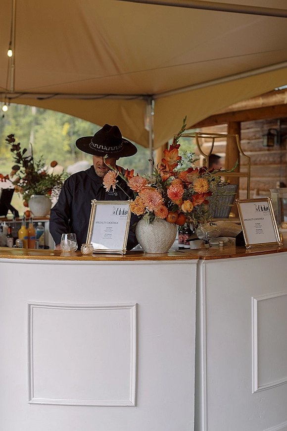 Wedding bar setup with a portable bar, framed cocktail menu signs, bottles and glassware under a tent with string lights and trees behind