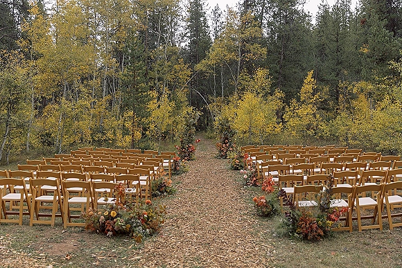 Ceremony setup with wood folding chairs lining a leaf-strewn aisle, dotted with greenery arrangements in an autumn forest grove
