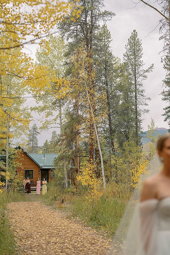 Processional moment as bride walks the aisle past bridesmaids waiting, her veil trailing on a gravel path through autumn forest by a cabin