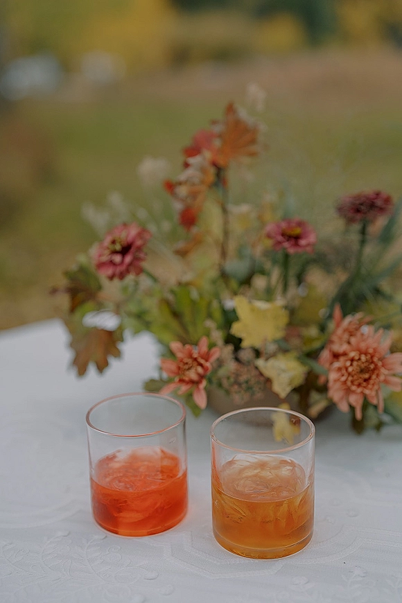 Wedding drinks and signature wedding cocktails in lowball glasses with ice beside a floral centerpiece on a white table outdoors