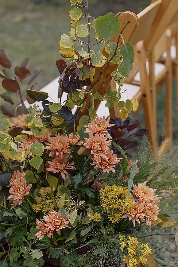 Ceremony chair flowers arranged on a wood folding chair with peach chrysanthemums, greenery, and yellow filler blooms on a grass lawn outdoors