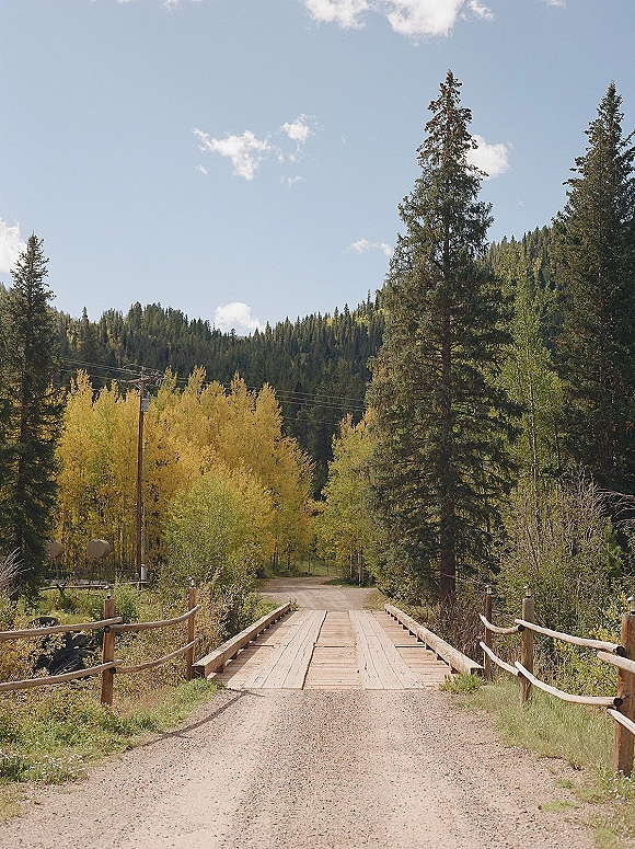 Outdoor wedding landscape with a wooden bridge and rustic fence leading to forest trees, mountain hillside, and blue sky with clouds