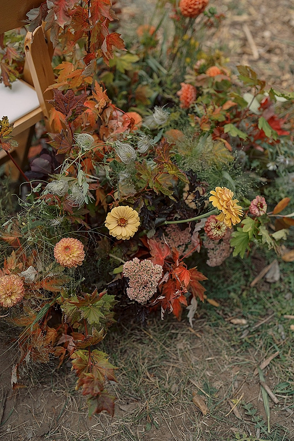 Wedding floral arrangement of fall wedding flowers with orange and yellow blooms, greenery, and dried seed pods beside a wooden chair on grass
