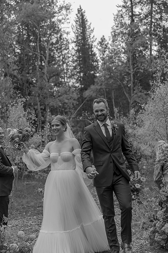 Wedding recessional of bride and groom walking hand in hand, bride holding bouquet and veil, smiling past guests on a forest path