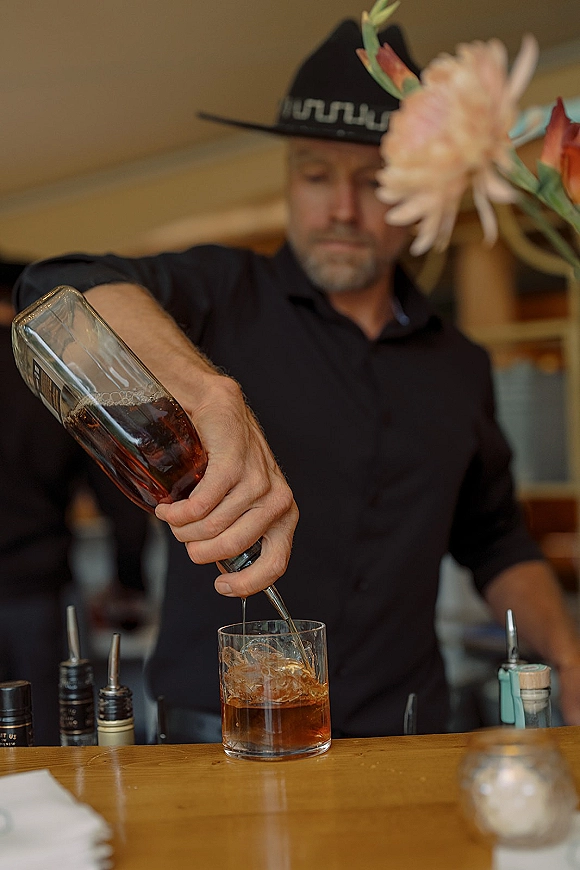 Wedding bar moment with a wedding bartender in a cowboy hat pouring whiskey into a rocks glass at an indoor bar with shelves behind