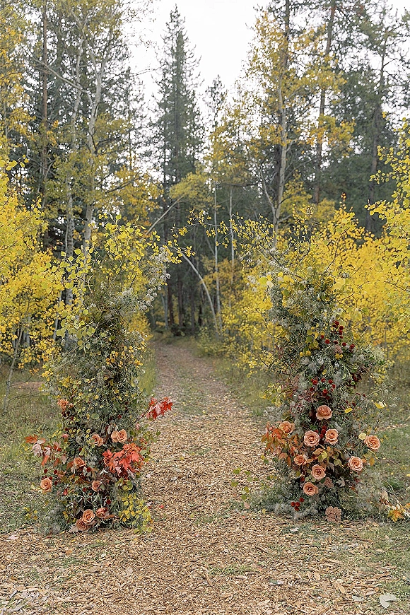 Ceremony aisle decor with rose and greenery arrangements, autumn leaves and berry stems lining a forest path with fall foliage