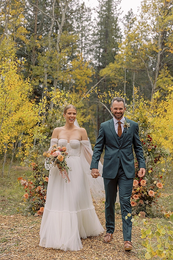 Wedding couple portrait of bride and groom walking hand in hand, bride in strapless dress and veil with bouquet on an autumn forest path