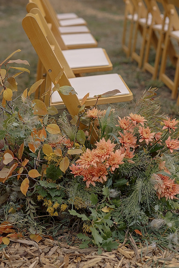 Ceremony aisle decor with an outdoor ceremony aisle of wood folding chairs, white cushions, and a ground floral arrangement on a grass lawn