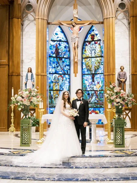 Couple portrait of the bride and groom at the church altar—a church wedding portrait framed by stained glass, her veil drapes toward marble floor.