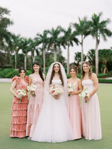 Bride and bridesmaids in blush bridesmaid dresses holding white bouquets on a palm-lined garden lawn beneath a softly cloudy sky.