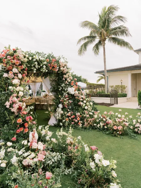 wedding floral arch frames a ceremony backdrop design with roses, set on a manicured lawn bordered by palm trees and open sky.