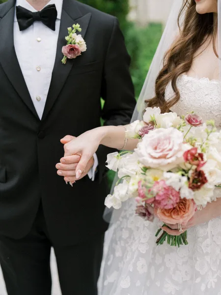 Bride and groom holding hands in a couple portrait, her bouquet resting between them, amid garden greenery and a stately stone building.