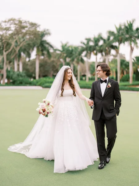 Couple portrait of a bride and groom walking among palm trees on a grass lawn, her veil trailing beneath an overcast sky.